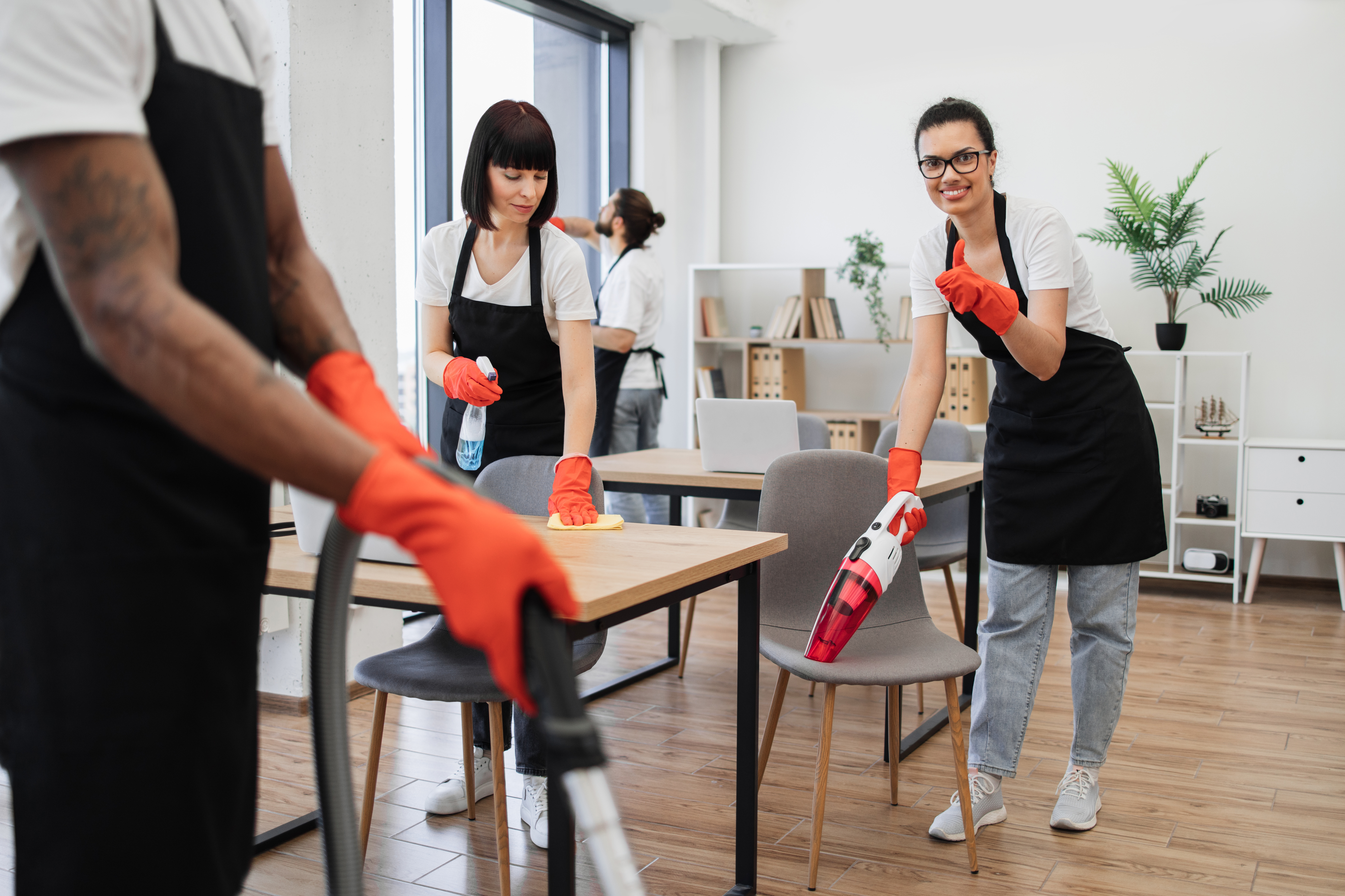 Multiethnic female cleaner vacuuming chair with portable vacuum cleaner.