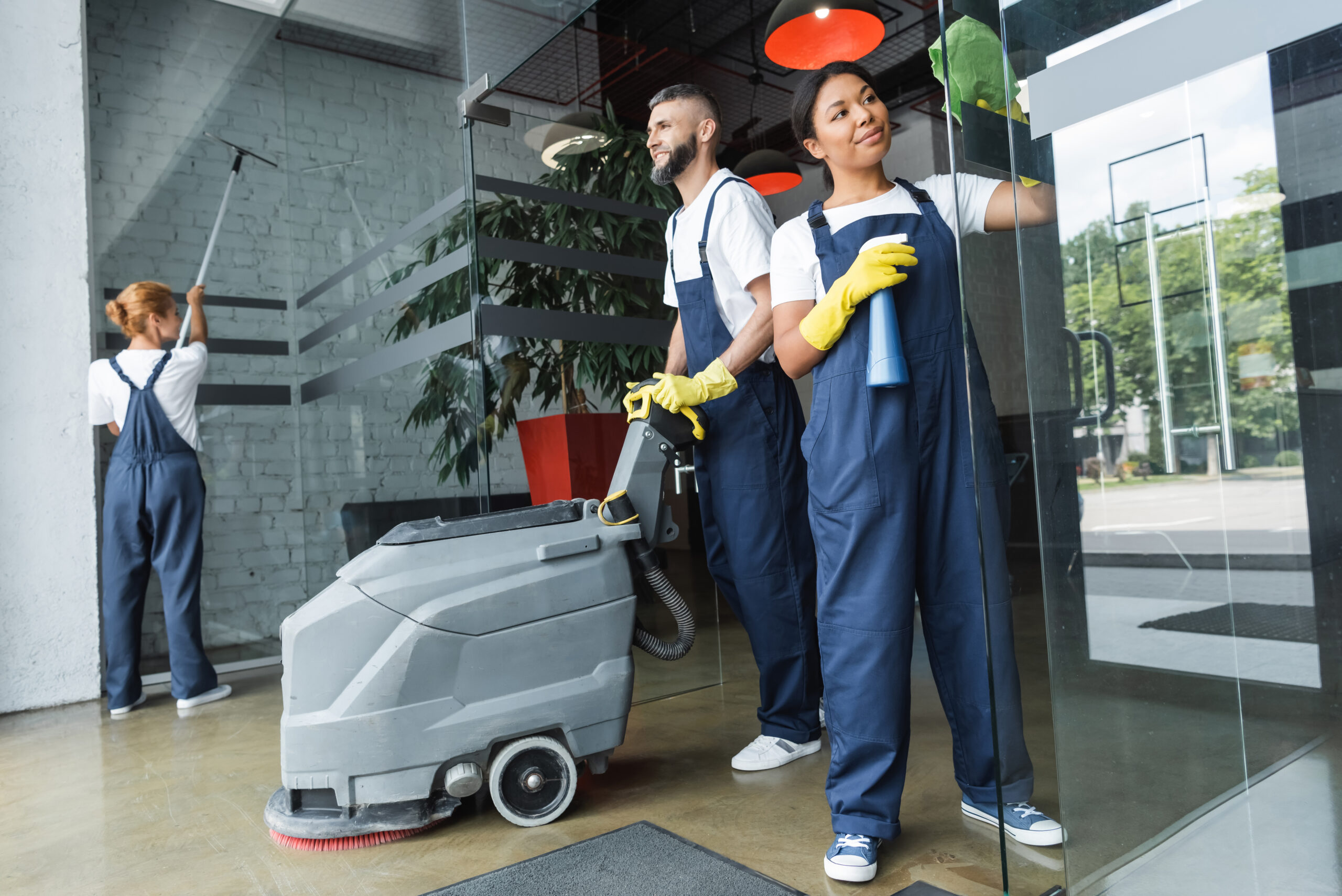 smiling man with floor scrubber machine near multiethnic women cleaning glass entrance of office