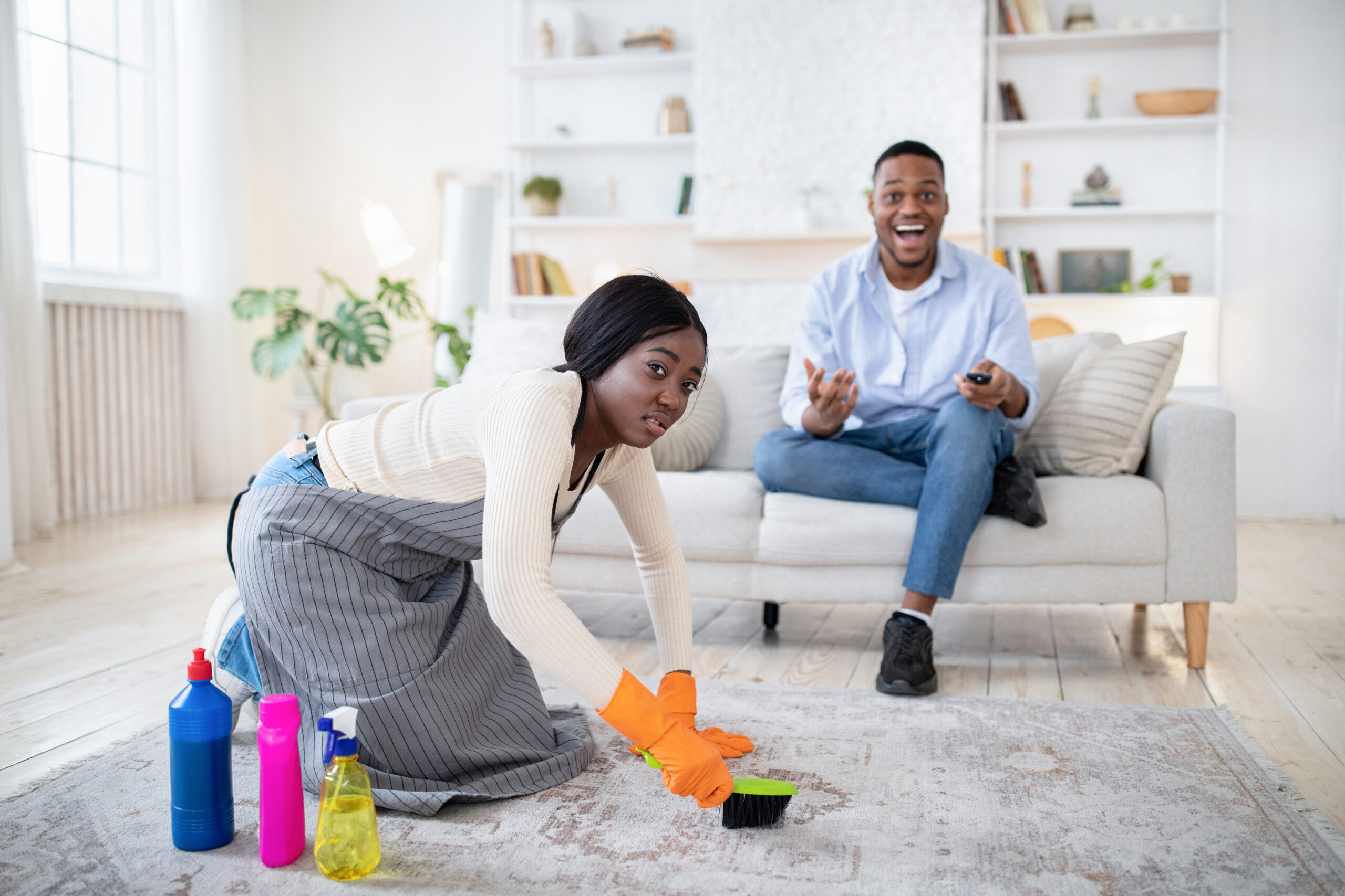 Young African American man watching television sitting on sofa, while his tired and offended wife cleaning house
